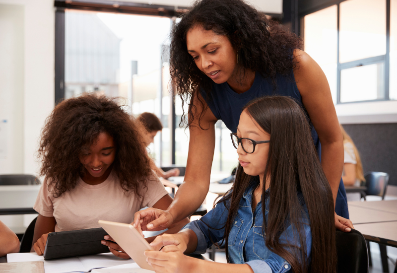 Teacher and students in classroom; teacher leans over toward two students working and gestures at book that one student holds
