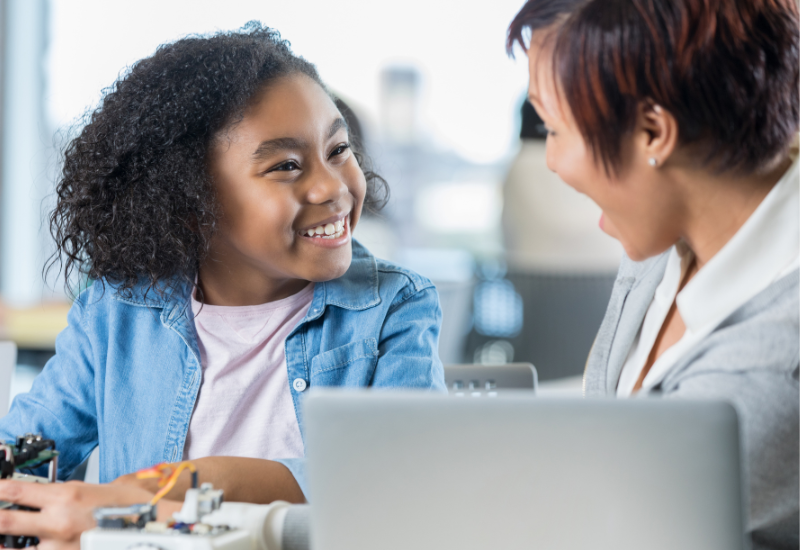 Student smiles at adult, sitting near laptop, at desk