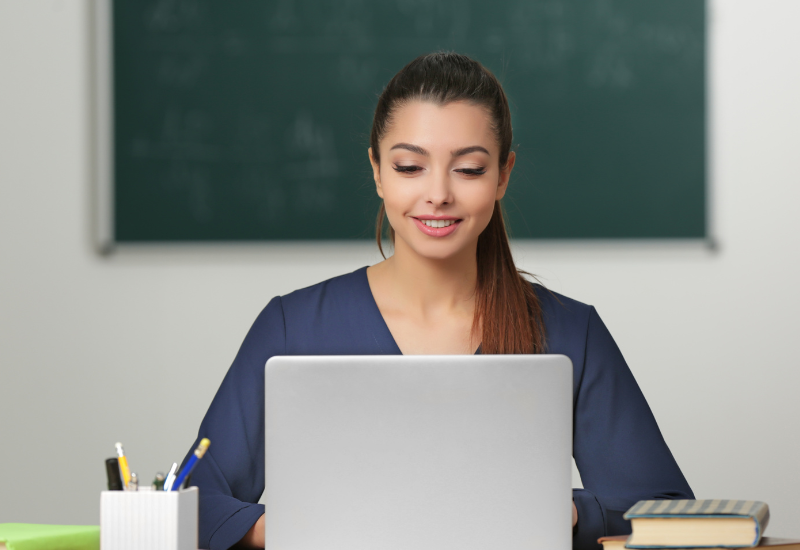 Teacher sits in classroom in front of blackboard, looking down at laptop