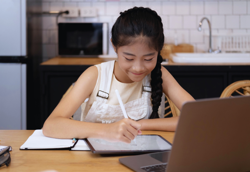 Student writes on tablet, at home in kitchen, in front of laptop