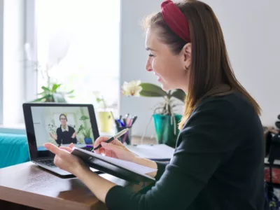 Student writing down in notebook while a teacher is on the screen of her laptop
