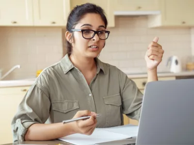 A woman sitting at a table with a laptop computer, gesturing to screen

