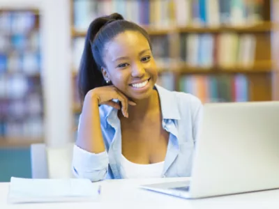 Girl sits in front of laptop inside library, smiling at camera
