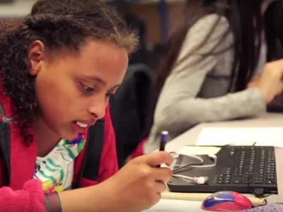 Girl writes on paper, sitting near computer
