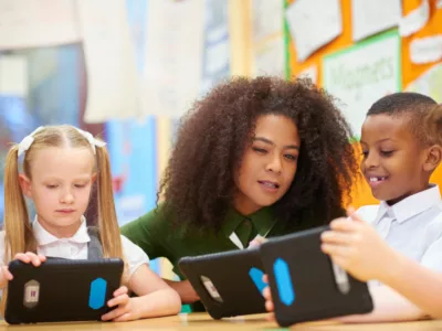 A group of children using a tablet computer in a classroom as teacher looks on
