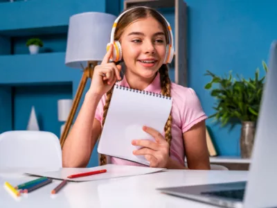 Student wearing headphones holds a notebook in hand in front of laptop
