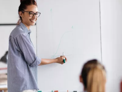Woman smiles, presenting at front of room with marker in hand
