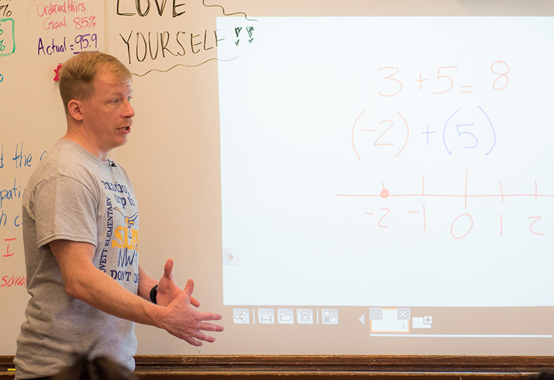 Teacher stands in front of whiteboard, looking out at class
