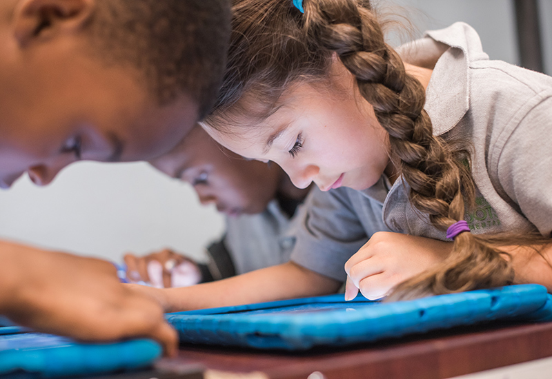 Focused image on one student looking down closely at tablet, with two other students in the background looking at tablets at the same table