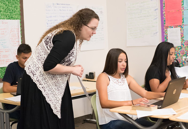Teacher stands behind student on laptop at desk, looking over student's shoulder as she speaks