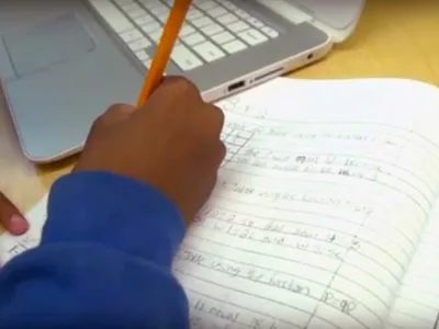 Photo closeup of student working in workbook with device closeby
