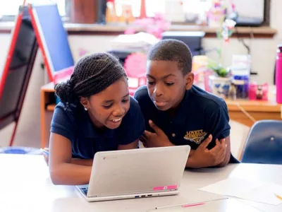 Two students work inside classroom, looking at laptop together
