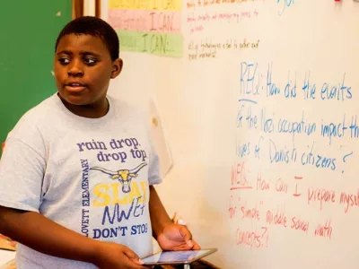 Student standing at white board with tablet in hand, looking back and speaking toward classroom
