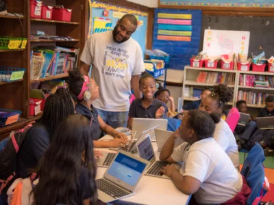 Teacher stands behind grouped desks of several students using laptops, smiling
