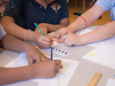 Close-up shot of several pairs of hands, holding pencils, writing on papers on desk
