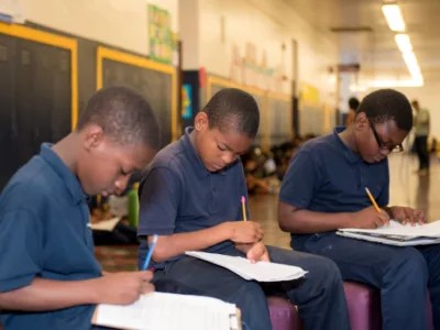 Three male students sitting in hallway, holding pencils and looking down at papers

