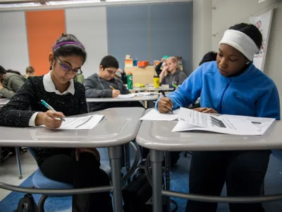 Two students complete work at their desks, sitting beside each other
