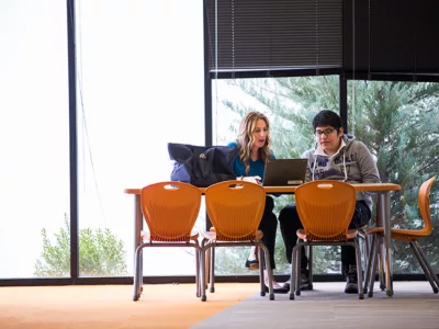 Teacher sits at table with student with windows in background, looking together at laptop
