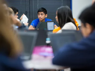 Students sit grouped at tables with technology
