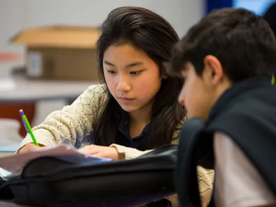 Close-up of a student working, with pencil in hand, on paper at desk, with blurred view of student to her side
