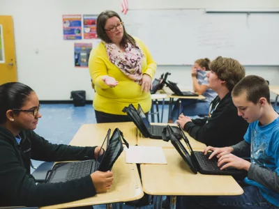 Teacher speaks with students seated at table
