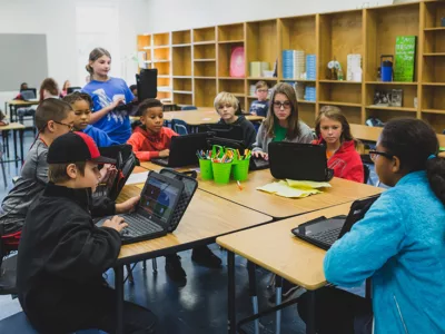 Students gather around a table, looking at one student at front of table

