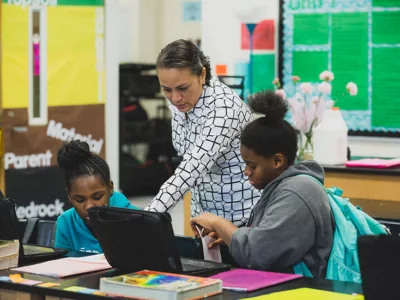 Teacher points at laptop while standing between two students
