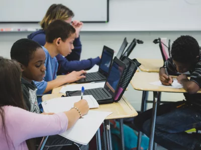 With pen in hand, teacher works with one student sitting in a group
