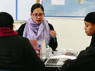 Teacher speaks, sitting at table with two students
