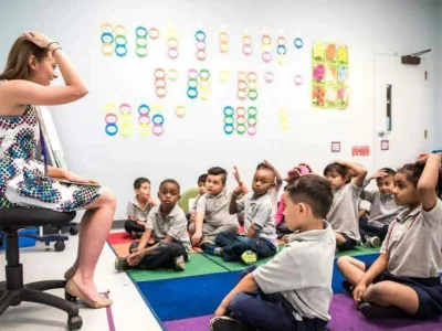 Photo of teacher seated at front of class with group of students seated in front and copying teacher patting head

