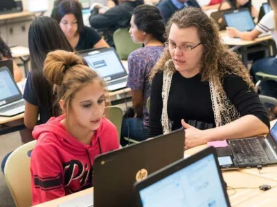 Photo of teacher and student in conversation seated at desk in front of laptop with other students in background

