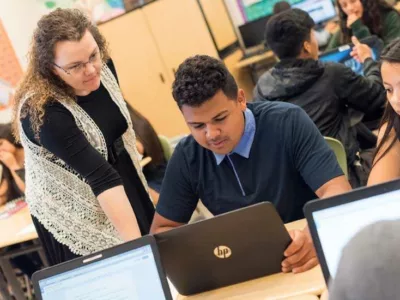 Photo of teacher standing and talking with student seated in front of his laptop
