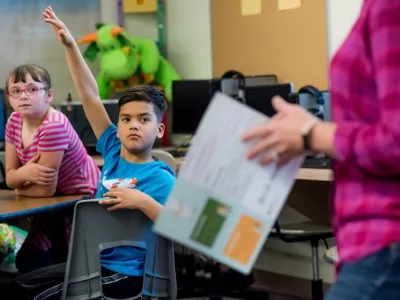 A seated student raises his hand, with blurred view of teacher in the foreground
