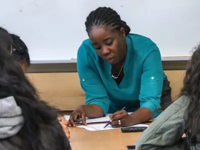 Teacher leans over desk, writing on paper, with students nearby
