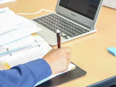 Photo of student arm writing in notebook with laptop in background
