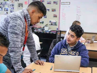 Photo of teacher aide standing and helping student with his work
