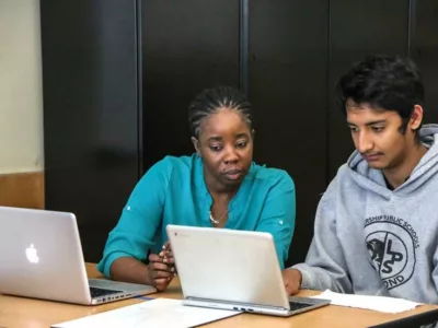 Photo of teacher and student working side by side at table with laptops directly in front of them
