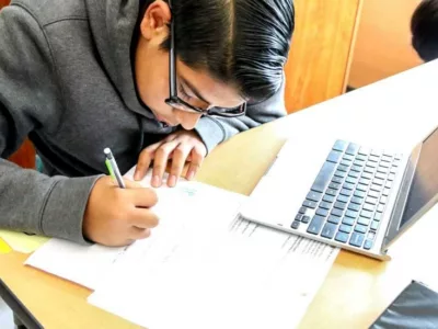 Photo of student working at table writing on worksheets with laptop in front of him
