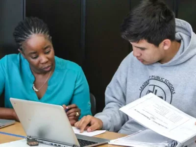 Photo of teacher and student working on a worksheet side by side at table with laptops directly in front of them
