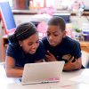 Two students work inside classroom, looking at laptop together